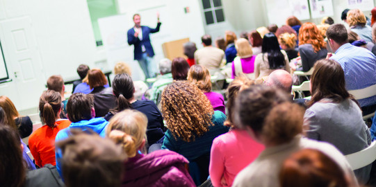 Veranstaltungen Publikum von hinten, im Hintergrund unscharf ein Vortragender auf einer Bühne